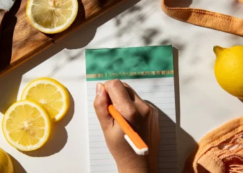 Flat-lay of fresh lemon slices on wooden board, green notepad with orange pen writing, yellow teapot, and orange mesh tote bag on marble surface.