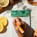 Flat-lay of fresh lemon slices on wooden board, green notepad with orange pen writing, yellow teapot, and orange mesh tote bag on marble surface.