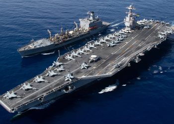 USS Gerald R. Ford aircraft carrier conducting underway replenishment at sea with supply ship alongside, fighter jets visible on flight deck.
