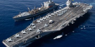 USS Gerald R. Ford aircraft carrier conducting underway replenishment at sea with supply ship alongside, fighter jets visible on flight deck.