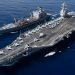 USS Gerald R. Ford aircraft carrier conducting underway replenishment at sea with supply ship alongside, fighter jets visible on flight deck.