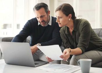 Middle‑aged couple sitting on a couch with a laptop and financial documents, reviewing their 2026 retirement planning and investment contributions at home.”