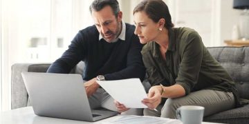 Middle‑aged couple sitting on a couch with a laptop and financial documents, reviewing their 2026 retirement planning and investment contributions at home.”