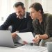 Middle‑aged couple sitting on a couch with a laptop and financial documents, reviewing their 2026 retirement planning and investment contributions at home.”
