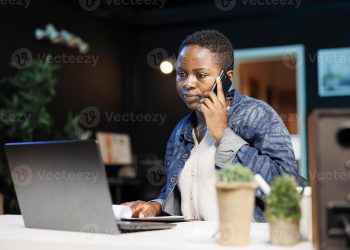Freelancer working from home office, using laptop and conversing with client on cell phone. Busy remote worker answering a call while comparing notes on wireless computer and notebook