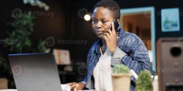 Freelancer working from home office, using laptop and conversing with client on cell phone. Busy remote worker answering a call while comparing notes on wireless computer and notebook