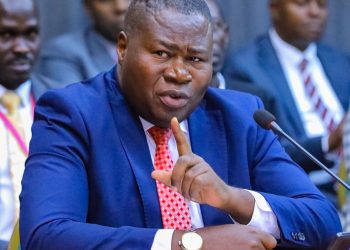 Ugandan politician speaking into a microphone during a parliamentary or committee session while gesturing with his finger in emphasis.