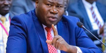Ugandan politician speaking into a microphone during a parliamentary or committee session while gesturing with his finger in emphasis.