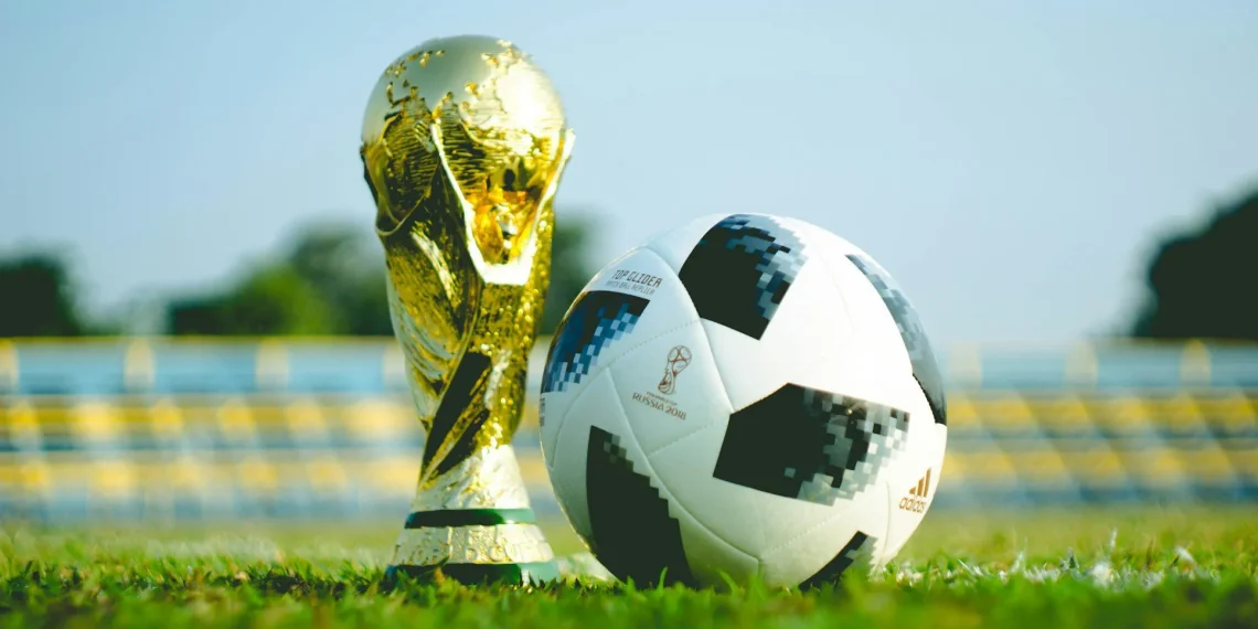 alt="FIFA World Cup Trophy and official match ball on grass in stadium"