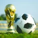 alt="FIFA World Cup Trophy and official match ball on grass in stadium"